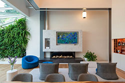 Modern living room featuring a Flare Double Corner 60” fireplace with an Invisible Mesh Safety Screen barrier, mounted below a wall-hung TV framed in marble, surrounded by contemporary seating, a vertical garden, and natural light.